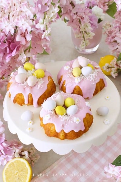 Three mini bundt cakes with pink icing are topped with pastel candy eggs and small white flowers, displayed on a white cake stand surrounded by pink flowers and a glass of pink drink—perfect for your Easter bundt cake recipe inspiration.