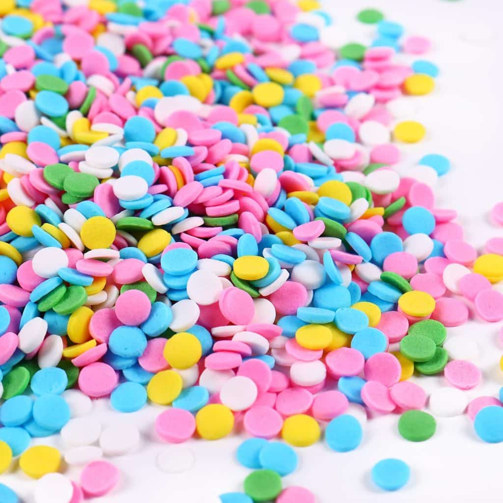 A colorful pile of round confetti sprinkles in pink, blue, yellow, green, and white scattered across a white surface—perfect for topping your favorite meringue cookies recipe.
