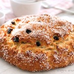 A round loaf of golden-brown bread topped with coarse sugar and raisins sits on a white surface, with white cups and a pink checkered cloth in the blurred background.
