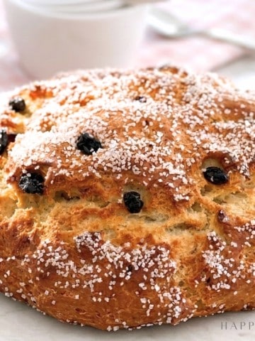 A round loaf of golden-brown bread topped with coarse sugar and raisins sits on a white surface, with white cups and a pink checkered cloth in the blurred background.