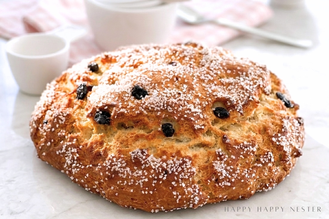 A round loaf of golden-brown bread topped with coarse sugar and raisins sits on a white surface, with white cups and a pink checkered cloth in the blurred background.