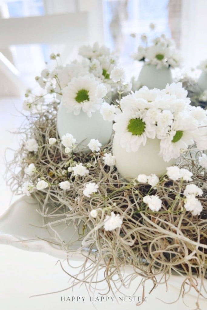 White flowers and green moss arranged in white egg-shaped vases, surrounded by delicate small white blooms, creating a soft, airy spring decor centerpiece on a white table.