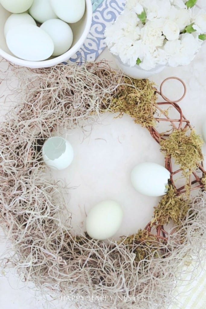 A partially completed spring flower wreath DIY with dried moss and twigs is surrounded by light blue eggs, some whole and one cracked, on a white surface with a bowl of eggs and white flowers nearby.