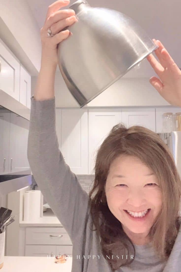 A smiling woman with long hair holds a large metal mixing bowl upside down over her head in a bright, modern kitchen.