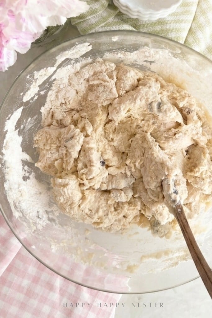 A glass bowl filled with partially mixed Irish soda bread dough (with buttermilk) and chocolate chips sits on a table. A wooden spoon rests in the bowl, flour coats the sides, and pink and green linens are underneath.