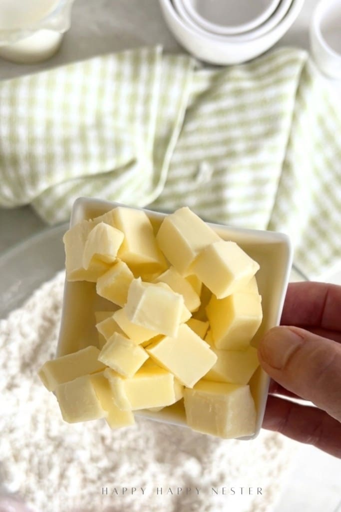 A hand holds a small white bowl filled with cubed butter above a bowl of flour, ready to make an Irish soda bread recipe (with buttermilk). A green and white striped cloth and measuring cups are in the background.