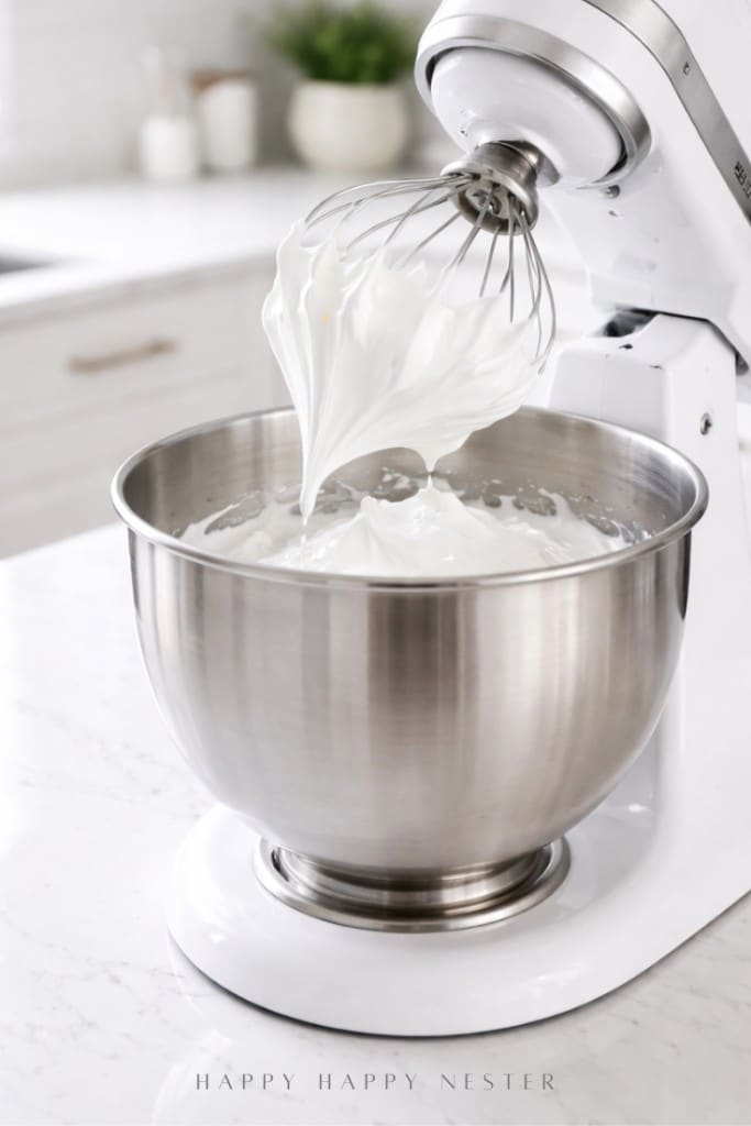 A stand mixer with a whisk attachment is whipping fluffy, white meringue in a stainless steel bowl on a white kitchen countertop.