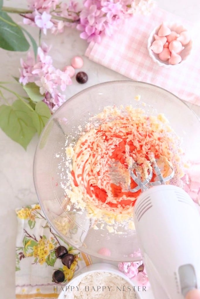 A hand mixer blends pink and white ingredients in a glass bowl on a floral-patterned cloth, surrounded by artificial flowers and a small bowl of pink meringues.