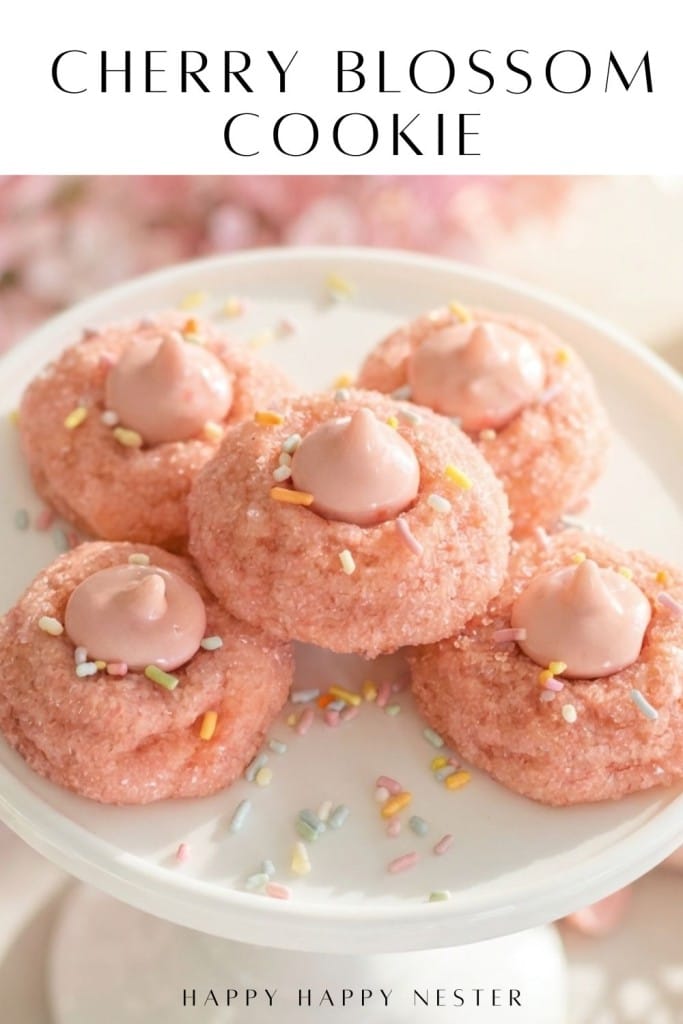 Five pink cherry blossom cookies with pink frosting centers and colorful sprinkles are displayed on a white cake stand, with “CHERRY BLOSSOM COOKIE” written above—perfect inspiration for your next cherry blossom cookie recipe.
