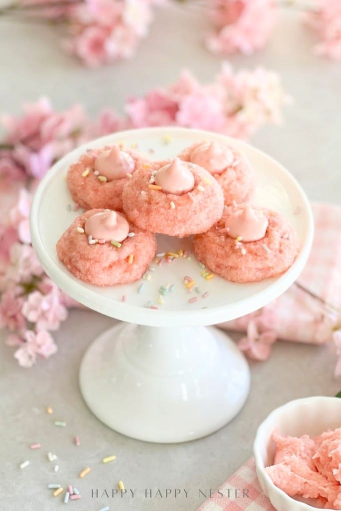 A white cake stand holds five pink cookies topped with pink frosting and colorful sprinkles. Pink flowers and a bowl of pink frosting surround the stand, creating a soft and festive display.