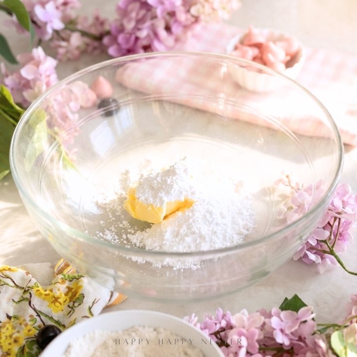 A glass mixing bowl with butter and powdered sugar inside, surrounded by pink flowers and a floral napkin on a light surface. A pink checkered cloth and small bowl are in the background.