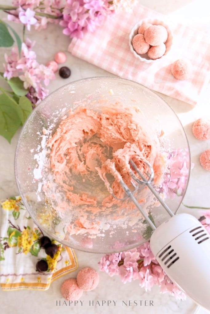 A bowl of pink frosting being mixed with a hand mixer, surrounded by pink flowers, cookies, and a floral napkin on a light surface.