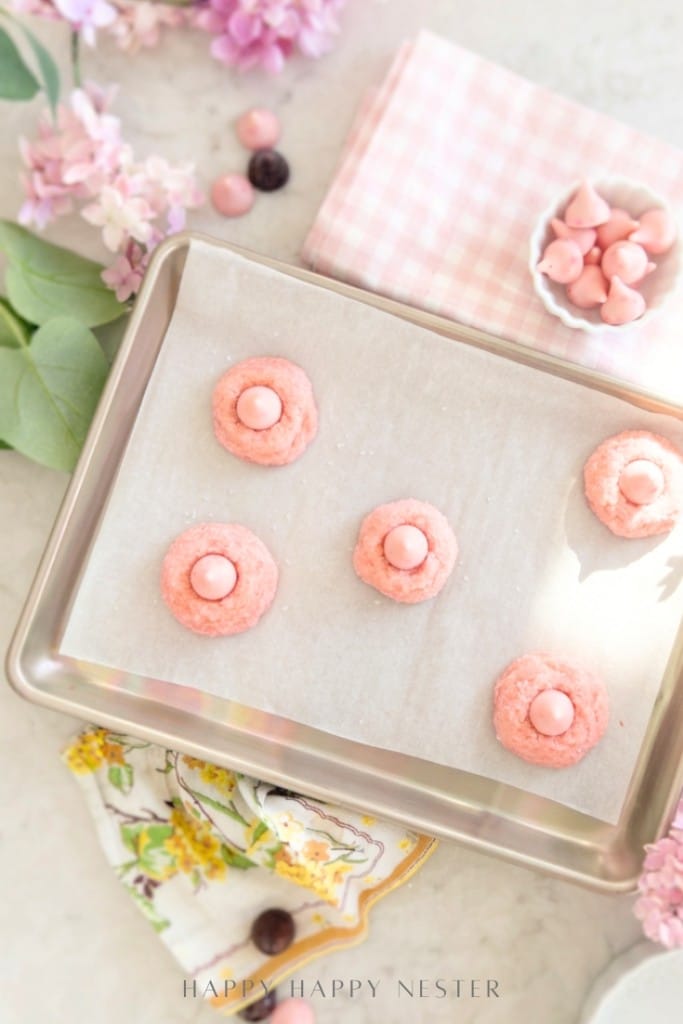 A baking sheet lined with parchment paper holds five unbaked pink cookies, each topped with a pink candy. Nearby are pink flowers, a floral napkin, a bowl of pink candies, and a pink gingham cloth.