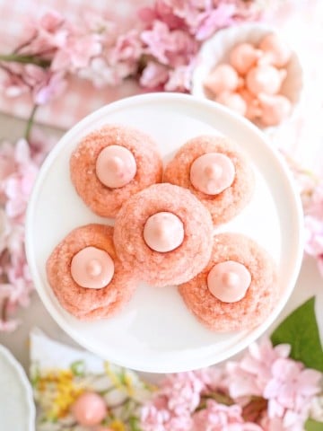 Five pink cookies with pink dollops in the center are arranged on a white plate, surrounded by pink cherry blossom flowers and pastel decorations, creating a soft, spring-themed scene.