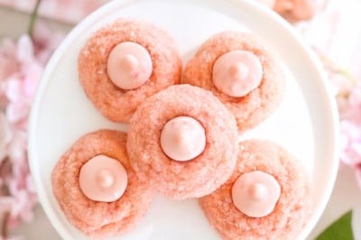 Five pink cookies with pink dollops in the center are arranged on a white plate, surrounded by pink cherry blossom flowers and pastel decorations, creating a soft, spring-themed scene.