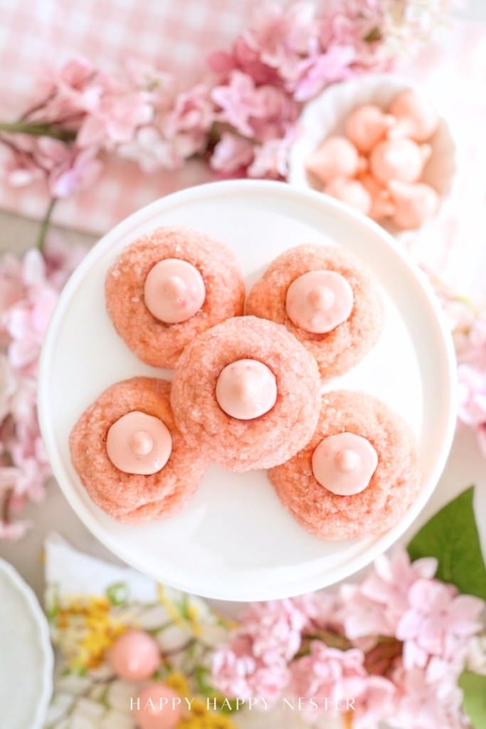 Five pink cookies with pink dollops in the center are arranged on a white plate, surrounded by pink cherry blossom flowers and pastel decorations, creating a soft, spring-themed scene.