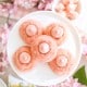 Five pink cookies with pink dollops in the center are arranged on a white plate, surrounded by pink cherry blossom flowers and pastel decorations, creating a soft, spring-themed scene.