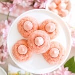 Five pink thumbprint cookies with pink frosting centers are arranged on a white plate, surrounded by pink flowers and a small bowl of round pink candies. The setting has a soft, springtime feel.