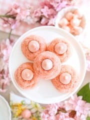 Five pink thumbprint cookies with pink frosting centers are arranged on a white plate, surrounded by pink flowers and a small bowl of round pink candies. The setting has a soft, springtime feel.