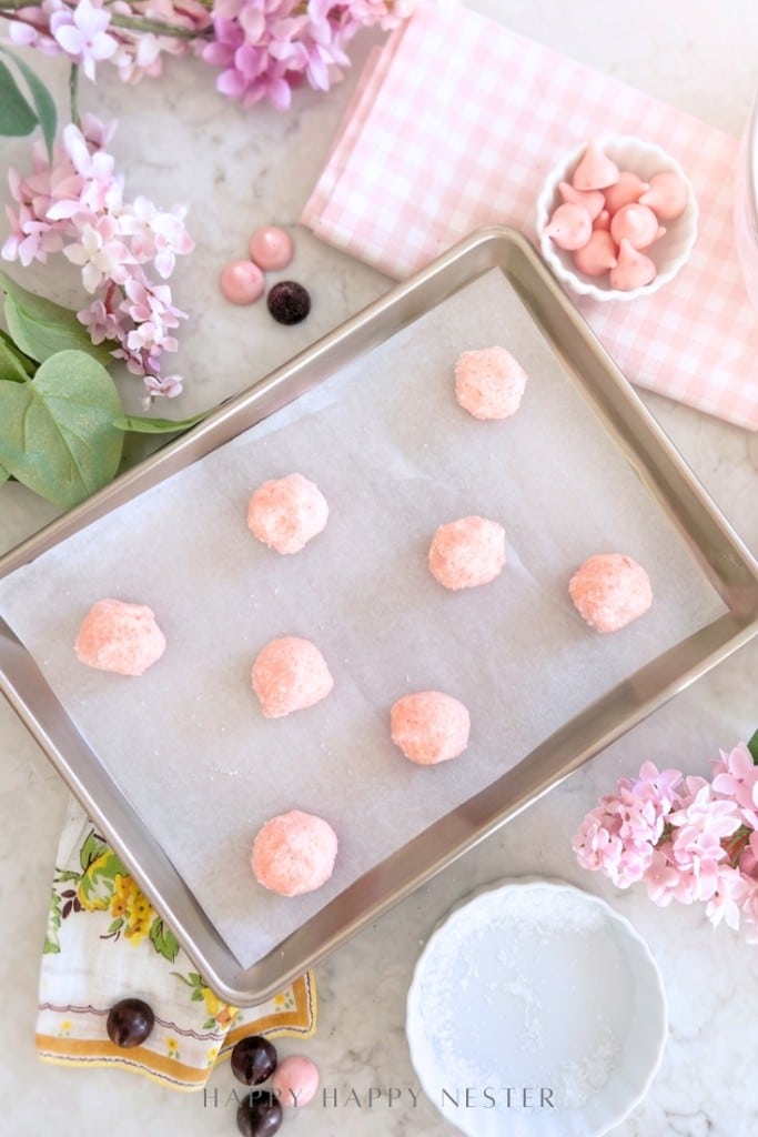 A baking tray lined with parchment paper holds eight pink dough balls. The tray is surrounded by lilac flowers, a pink cloth, pink candies, and a small bowl of white powder on a marble surface.