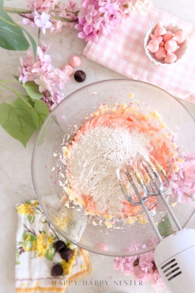 A glass bowl with pink cookie dough from a cherry blossom cookie recipe is mixed by a hand mixer on a marble surface, surrounded by pink flowers, a floral napkin, and a small dish of pink candies.