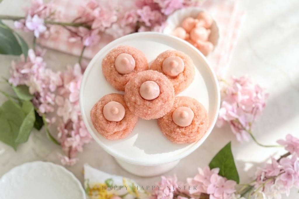 Five pink cookies with pink frosting centers, inspired by a cherry blossom cookie recipe, are arranged in a flower shape on a white cake stand, surrounded by pink flowers and soft pastel decor.