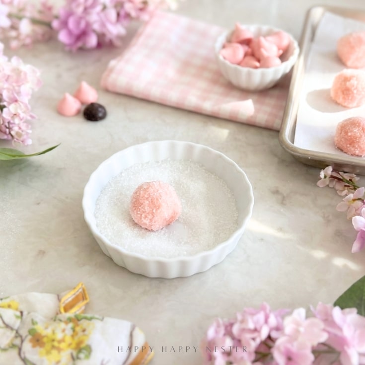 A pink truffle is being rolled in sugar in a white dish, surrounded by more pink truffles on a baking tray, lilac flowers, a checked cloth, and a floral napkin on a light countertop.