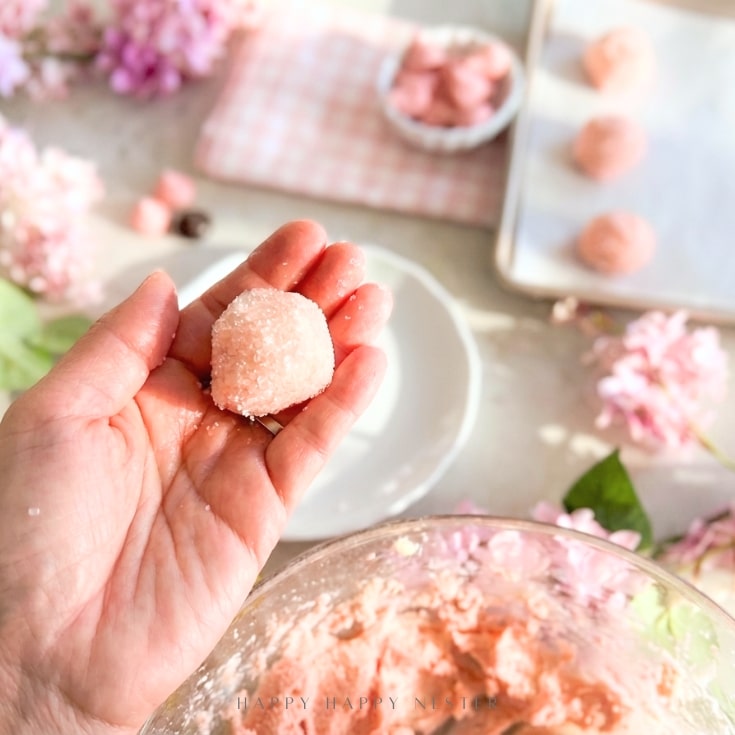 A hand holds a pink sugar-coated ball of dough, ready for baking cherry blossom cookie recipe treats, with more dough balls on a tray and pink flowers in the background, evoking a charming springtime baking scene.