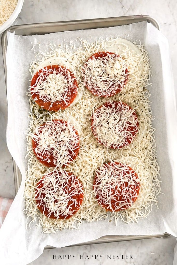 A parchment-lined baking sheet holds six sliced tomatoes topped with shredded cheese, surrounded by more shredded cheese on the sheet. The scene is on a light countertop.