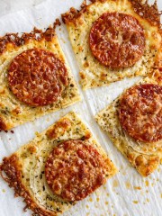 Four square slices of homemade onion ring pizza recipe on parchment paper, each topped with melted cheese, herbs, onions, a slice of pepperoni, and crispy edges where the cheese is browned.