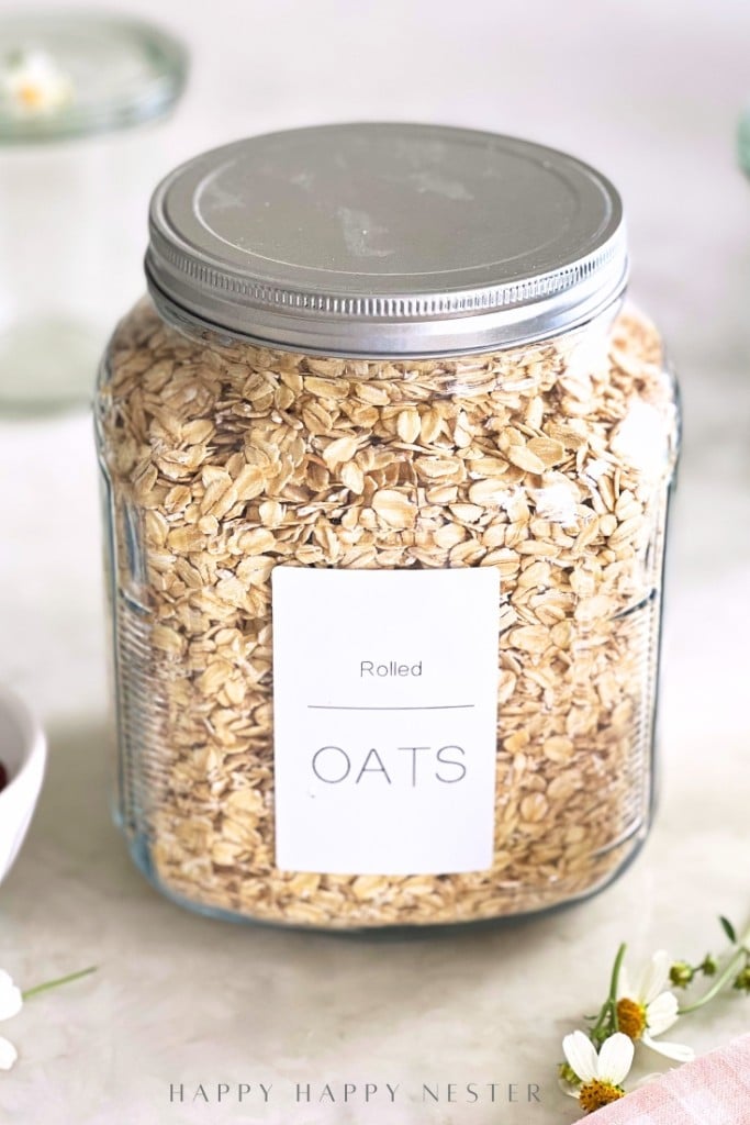 A glass jar filled with rolled oats, labeled "Rolled OATS" with a metal lid, sits on a white surface—perfect for trying an overnight oats recipe with yogurt. The background includes soft-focus jars and small white flowers.