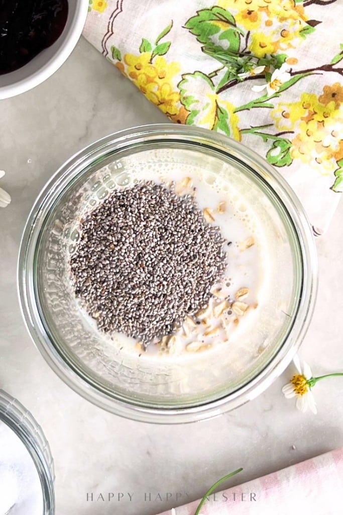 A glass jar filled with oats, milk, and a layer of chia seeds sits on a light surface near a yellow-flowered napkin.