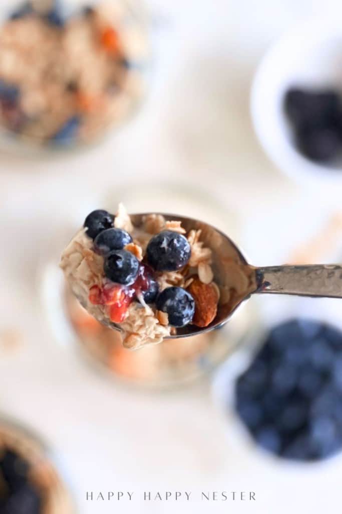 A close-up of a spoon holding oatmeal topped with blueberries, granola, and nuts, with bowls of blueberries and oatmeal blurred in the background.
