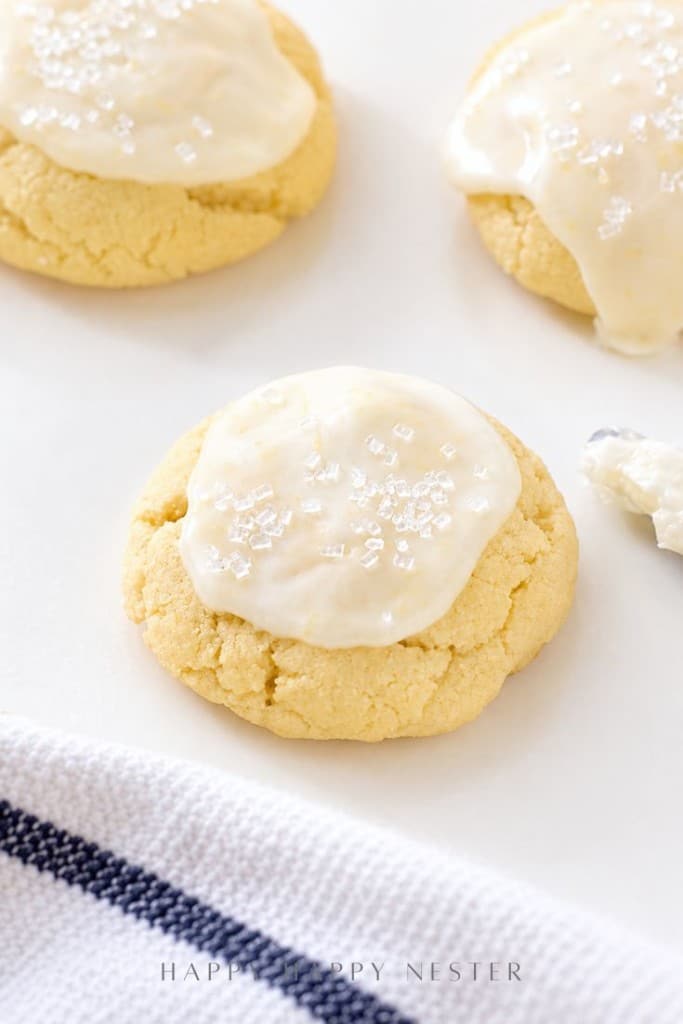 Three soft sugar cookies topped with white icing and sparkling sugar crystals are arranged on a white surface, showcasing a classic lemon sugar cookie recipe. A striped towel and an icing knife are partially visible nearby.