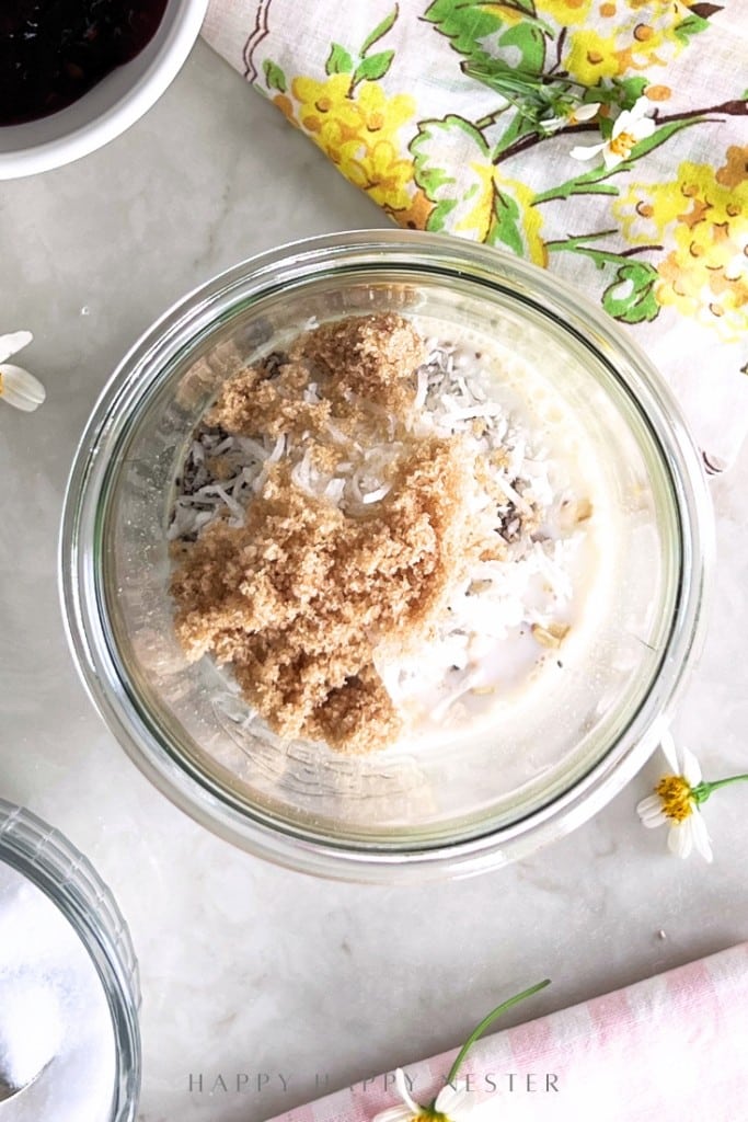 A glass bowl filled with oats, shredded coconut, brown sugar, and milk sits on a marble surface near a floral napkin, a white bowl, and small white flowers.