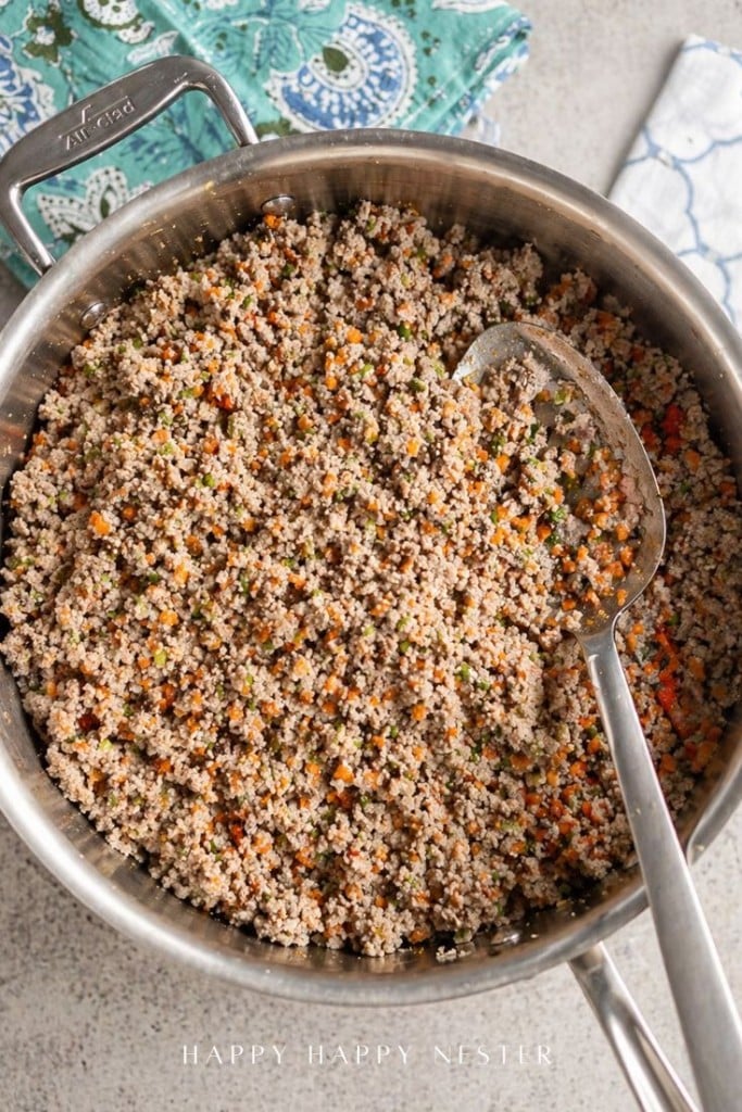 A stainless steel pot filled with cooked ground meat mixed with finely chopped vegetables, including carrots and herbs—perfect as a savory filling for your favorite fried wonton recipe. A large metal spoon rests inside the pot.
