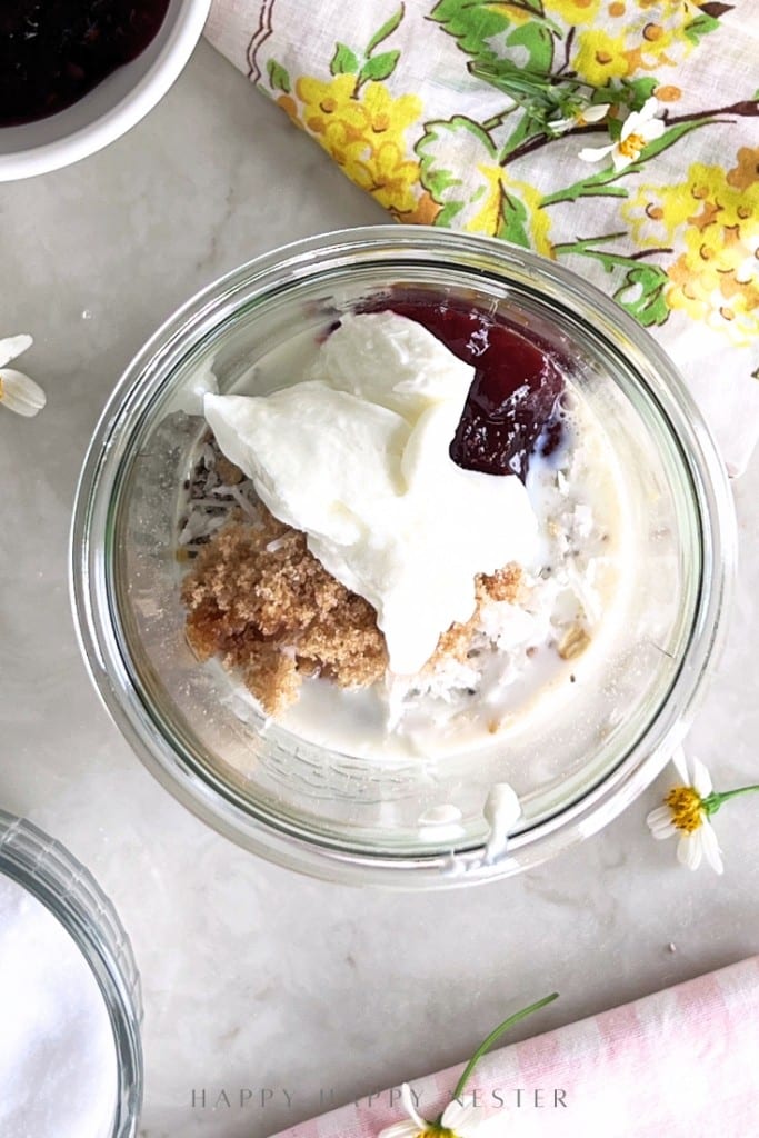 A glass jar filled with milk, brown sugar, whipped cream, and a dollop of red jam sits on a light surface, perfect for an overnight oats recipe with yogurt. A floral napkin, small flowers, and bowls with other ingredients are nearby.