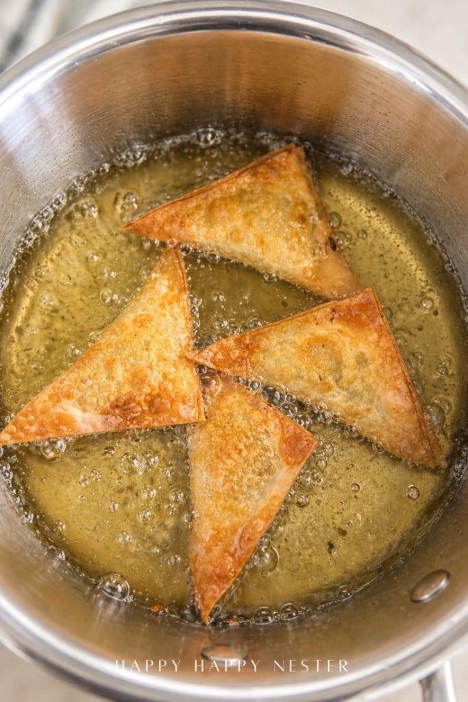 Four golden-brown triangular wontons are frying in hot oil inside a stainless steel pot, bubbles forming around them as they cook—an irresistible scene straight from a classic fried wonton recipe.