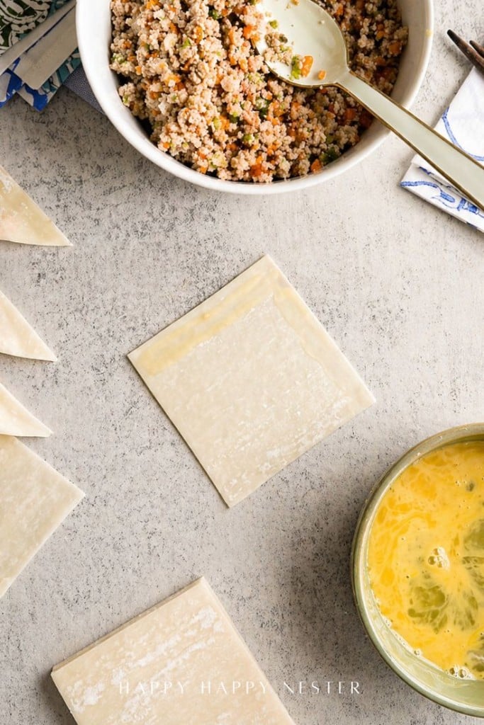Wonton wrappers on a countertop, one brushed with egg wash, next to a bowl of beaten eggs and a bowl of quinoa salad. A spoon rests in the salad.