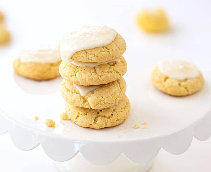 A stack of three frosted cookies sits on a white cake stand, with additional cookies in the background. The cookies are pale yellow and topped with a glossy white icing.
