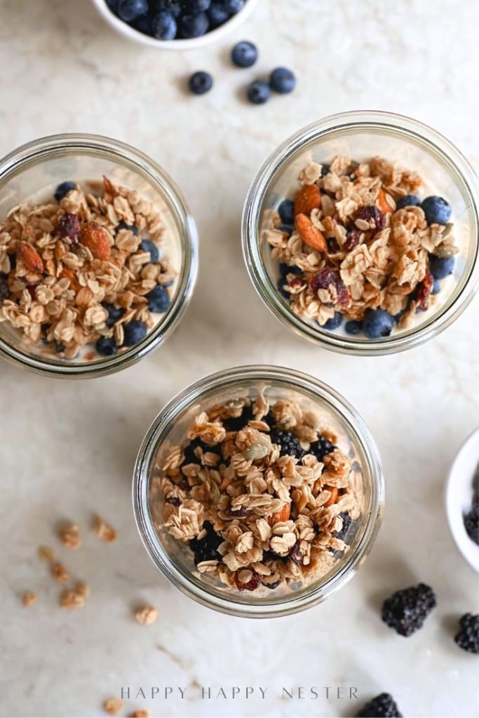 Three glass jars filled with an overnight oats recipe with yogurt, granola, nuts, and fresh berries sit on a light countertop. Scattered granola pieces and bowls of blueberries and blackberries are visible around the jars.
