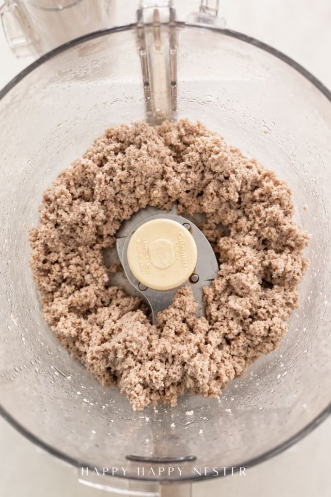 Ground nut mixture in a food processor bowl with metal blades, seen from above. The mixture appears finely processed and slightly coarse, with the lid partially visible at the top.