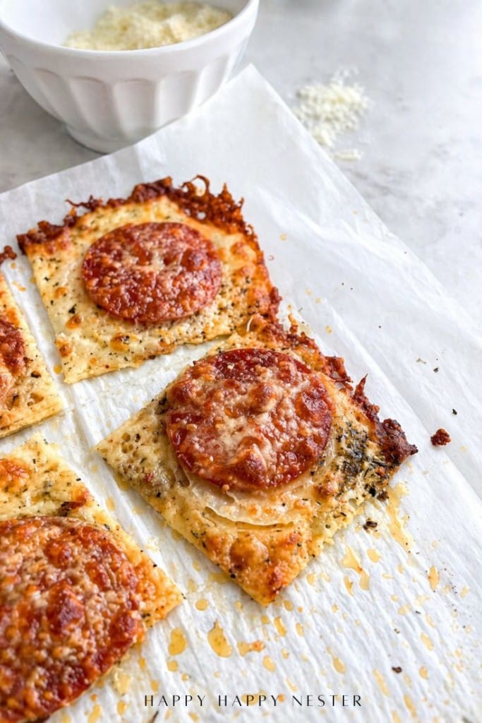 Close-up of crispy, square pepperoni pizza slices with melted cheese and herbs on parchment paper. A white bowl with grated cheese is in the background, perfect inspiration for trying a new onion ring pizza recipe.