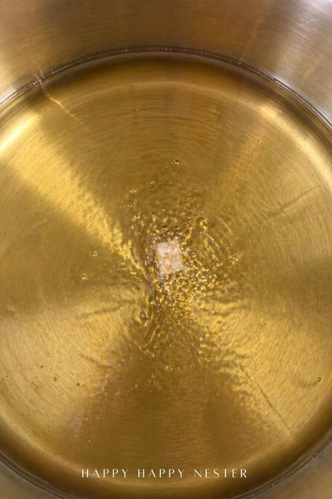 A single sugar cube is dissolving in hot water inside a shiny, gold-colored metal pot. The text "HAPPY HAPPY NESTER" is written at the bottom of the image.