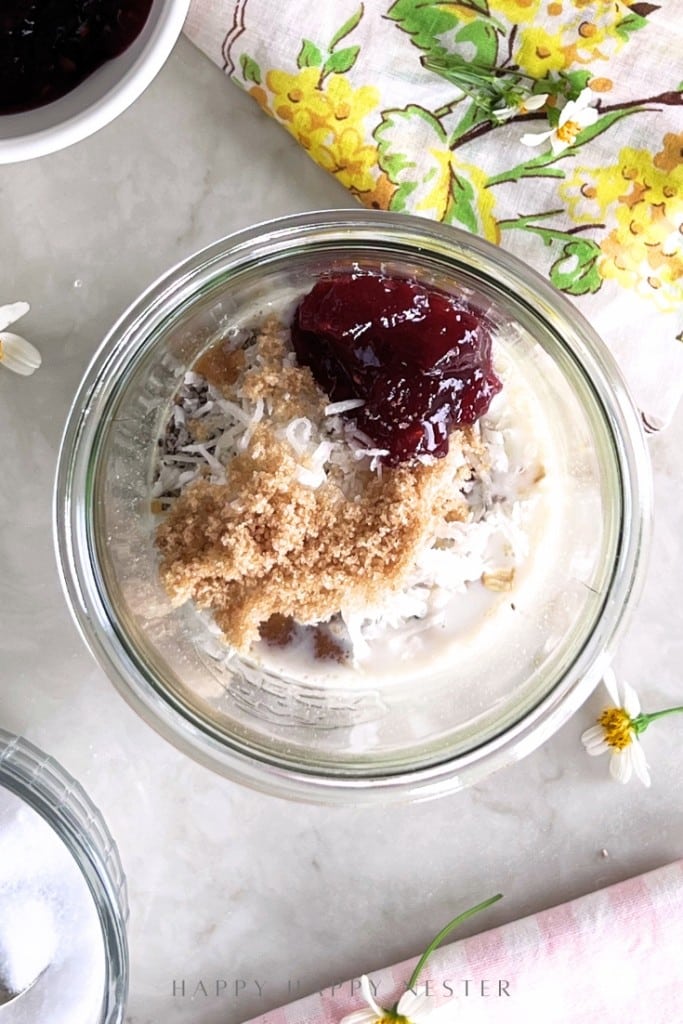 A glass jar filled with oats, brown sugar, shredded coconut, milk, yogurt, and a dollop of berry jam sits on a marble surface beside a floral napkin and small white flowers—an inviting overnight oats recipe with yogurt.