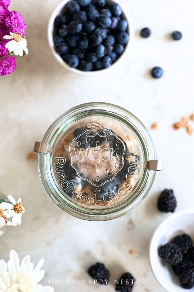Overhead view of a glass Weck jar with a lid, surrounded by bowls of blueberries and blackberries, scattered berries, and white and purple flowers on a light surface.