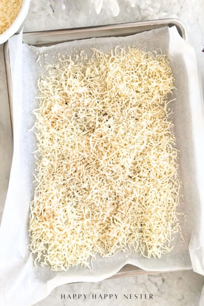 A baking sheet lined with parchment paper is covered with shredded cheese and some seasoning, ready to be baked.