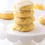 A stack of four soft, yellow cookies made from a delicious lemon sugar cookie recipe with white icing and sprinkles sits on a white scalloped cake stand. More cookies are visible in the background, all in a bright, light-filled setting.