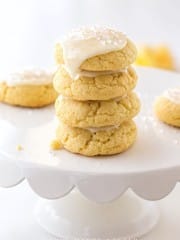 A stack of four soft, yellow cookies made from a delicious lemon sugar cookie recipe with white icing and sprinkles sits on a white scalloped cake stand. More cookies are visible in the background, all in a bright, light-filled setting.