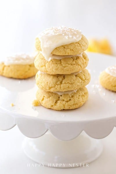 A stack of four soft, yellow cookies made from a delicious lemon sugar cookie recipe with white icing and sprinkles sits on a white scalloped cake stand. More cookies are visible in the background, all in a bright, light-filled setting.